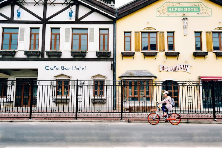 An Asian woman cycling along a street on a bicycle. The bicycle looks like one that's part of a public bicycle hire scheme. In the background are two buildings, 'Cafe bar hotel' and 'Alpen hotel restaurant', both have similar Alpen architecture. The main difference between the two is the cafe is painted white, whereas the restaurant is painted light yellow